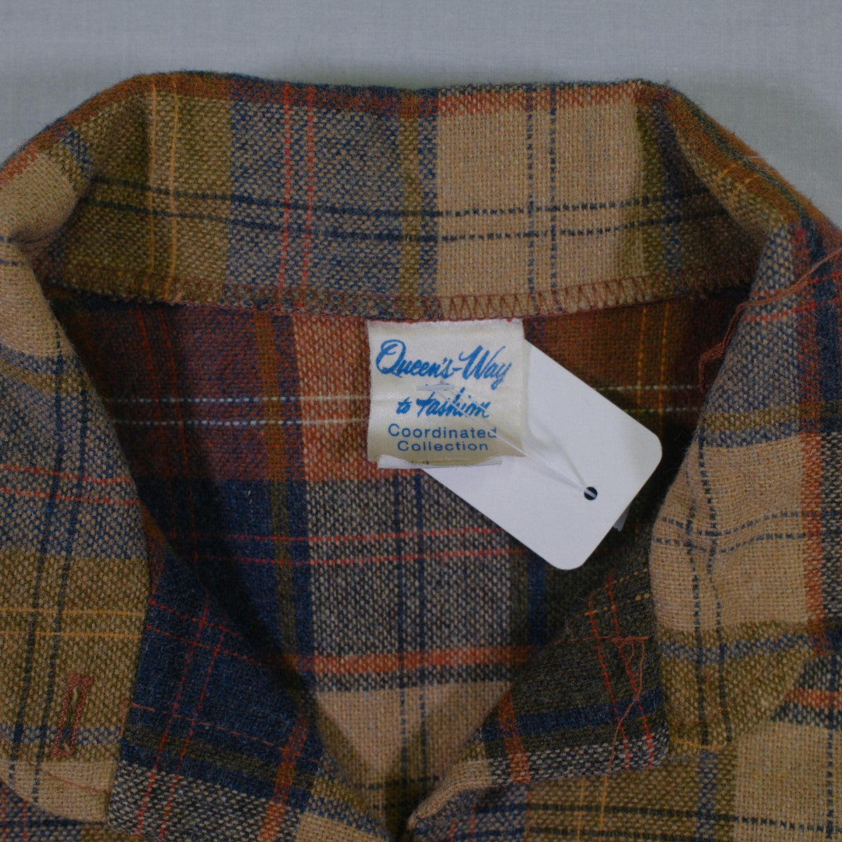 A close-up of a plaid button-down shirt collar in a tan, brown, and navy tartan pattern, with a visible 'Queen's Way Coordinated Collection' paper tag.